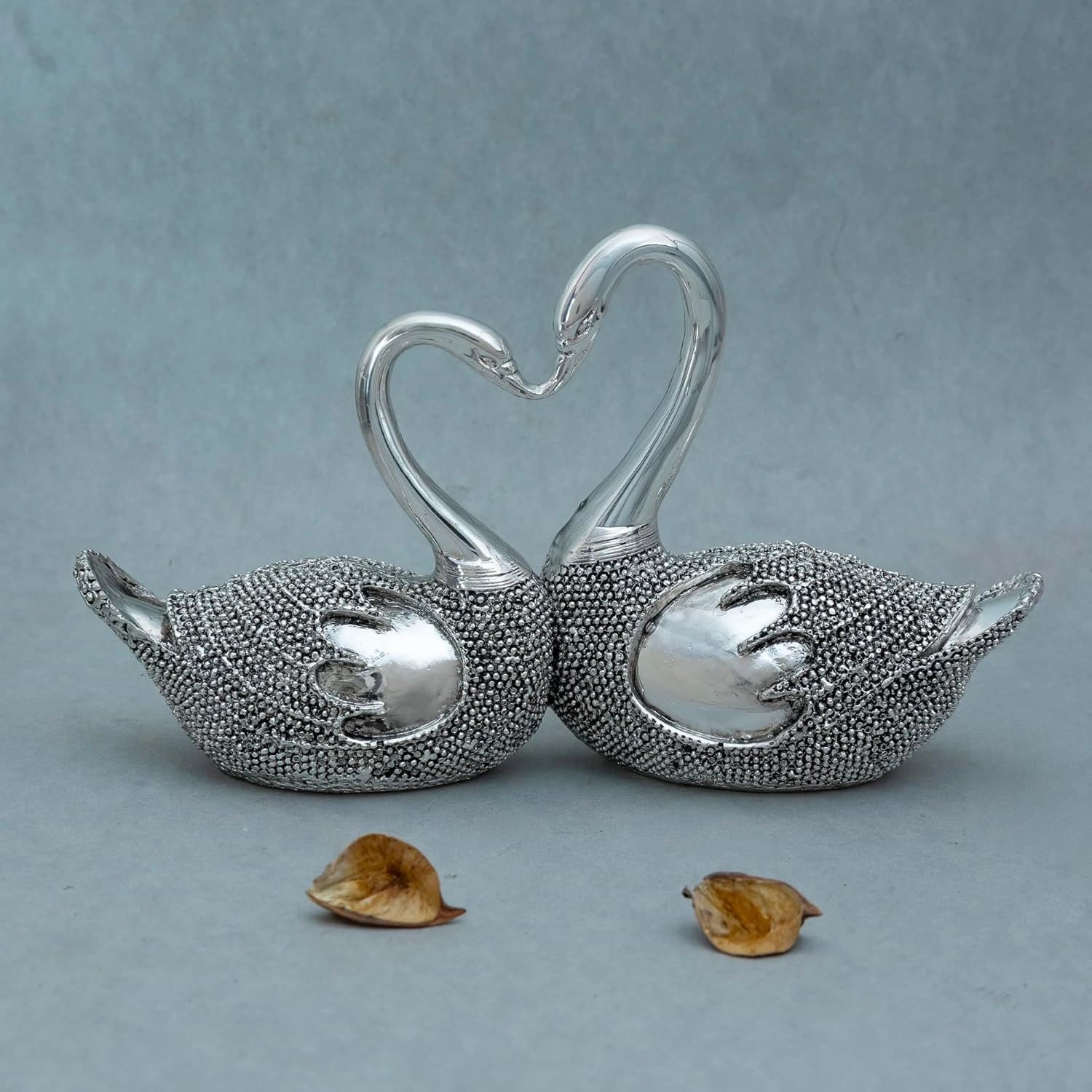 Silver decorative lovebird sculpture with textured bodies and entwined necks forming a heart shape on grey background