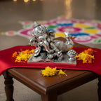 Silver Lord Ganesha idol reclining on a book with bowl of sweets on wooden table with yellow marigold flowers