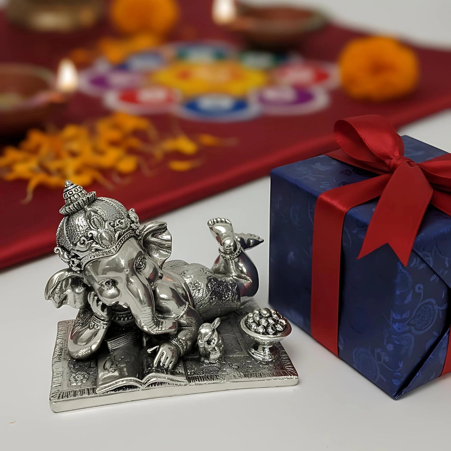Silver Ganesh idol reading book with blue gift box and red ribbon on white surface