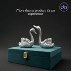Two polished silver swan figurines on a teal decorative box with gold clasp against dark background