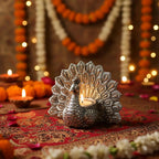 Silver peacock tealight candle holder on a red patterned cloth with marigold garlands and diyas in the background