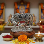 Silver cow and calf statue on wooden base with marigold flowers, red powder bowl, and lit brass oil lamp