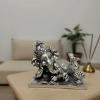 Silver Lord Ganesha statue reclining on a book on a wooden table with blurred sofa and plant background