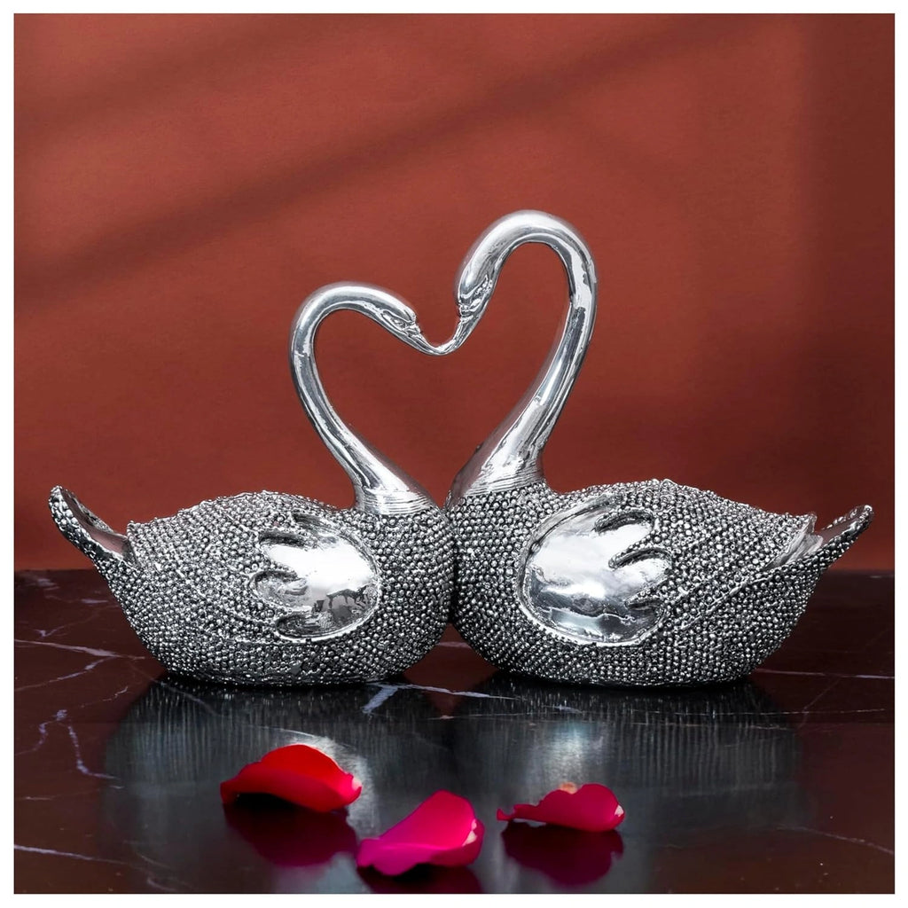 Pair of silver swan figurines with textured bodies on a dark surface with red rose petals