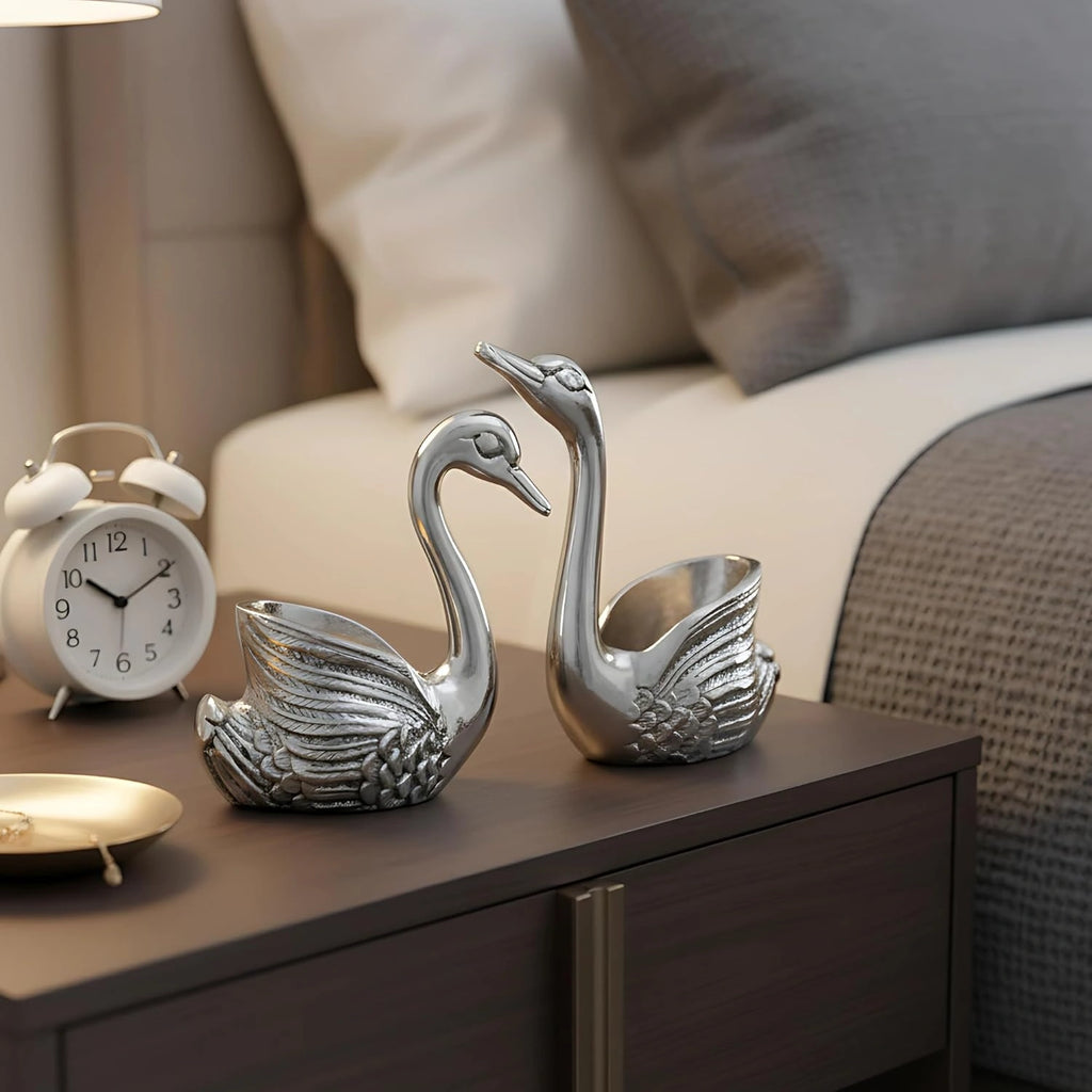Silver swan figurines on a dark wooden bedside table with white alarm clock and bed background
