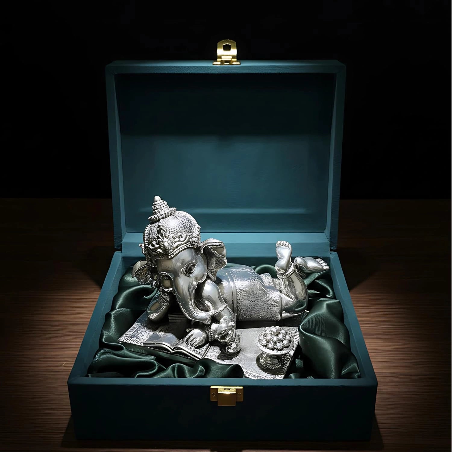 Silver Ganesh idol lying on its stomach reading a book, placed in a green velvet-lined blue gift box