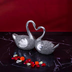 Decorative silver swan figurines with curved necks forming a heart, placed on black marble with red and white flower petals
