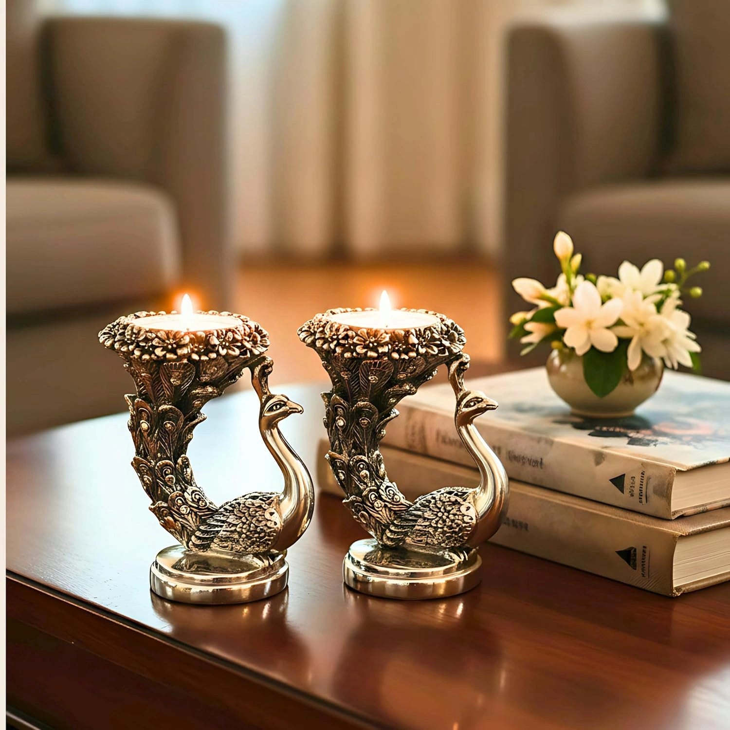 Pair of ornate brass peacock candle holders with lit candles on wooden table, next to books and white flowers