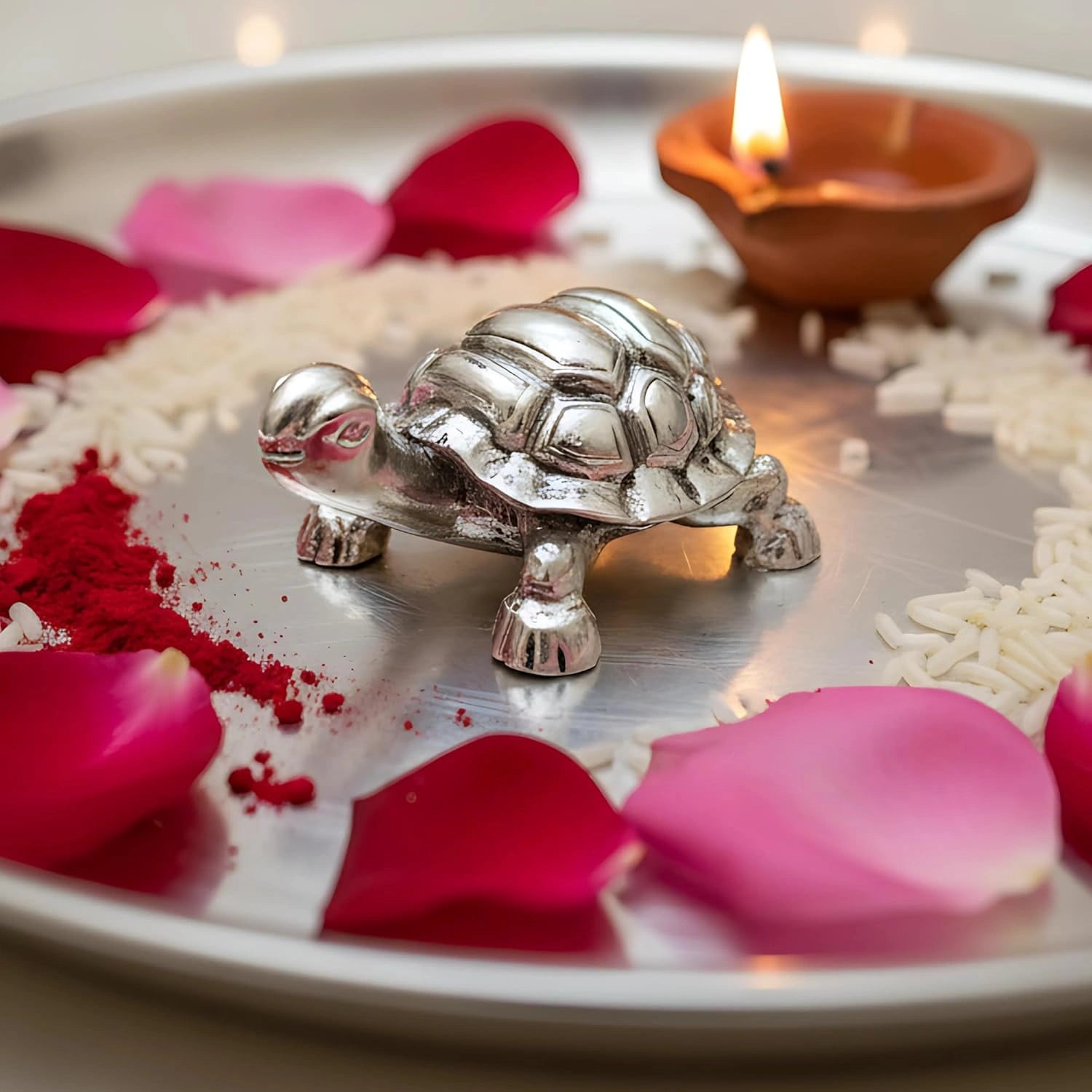 Silver turtle figurine on metal plate decorated with rose petals, red powder, rice, and a lit diya lamp