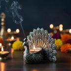 Silver peacock-shaped diya holder with lit tealight candle and smoke, festive floral background