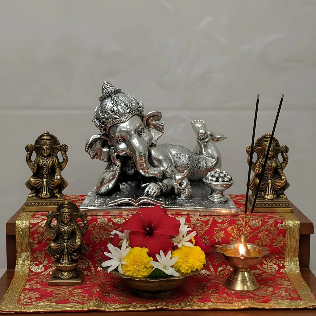 Silver reclining Lord Ganesha idol with brass Lakshmi statues, brass diya lamp, incense sticks, and fresh flowers on red cloth