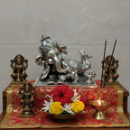 Silver reclining Lord Ganesha idol with brass Lakshmi statues, brass diya lamp, incense sticks, and fresh flowers on red cloth
