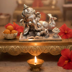 Silver Ganesha idol in relaxed pose on ornate gold tray with red hibiscus flowers, sweets, and lit oil lamp