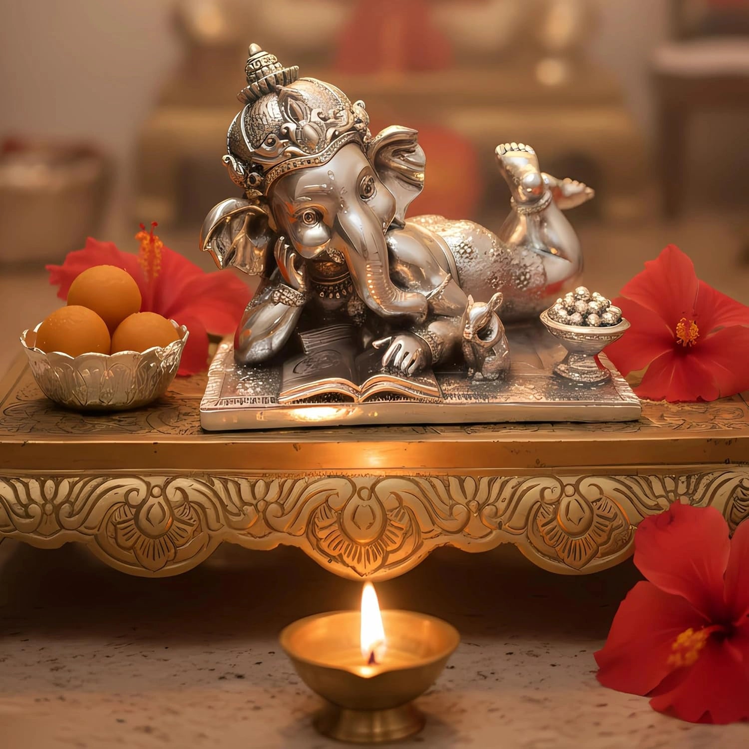 Silver Ganesha idol in relaxed pose on ornate gold tray with red hibiscus flowers, sweets, and lit oil lamp