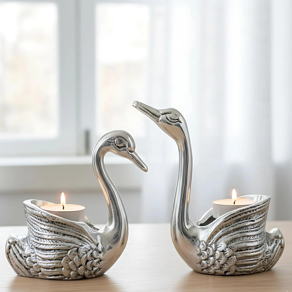 Pair of silver swan-shaped candle holders with lit tea candles on wooden table near window