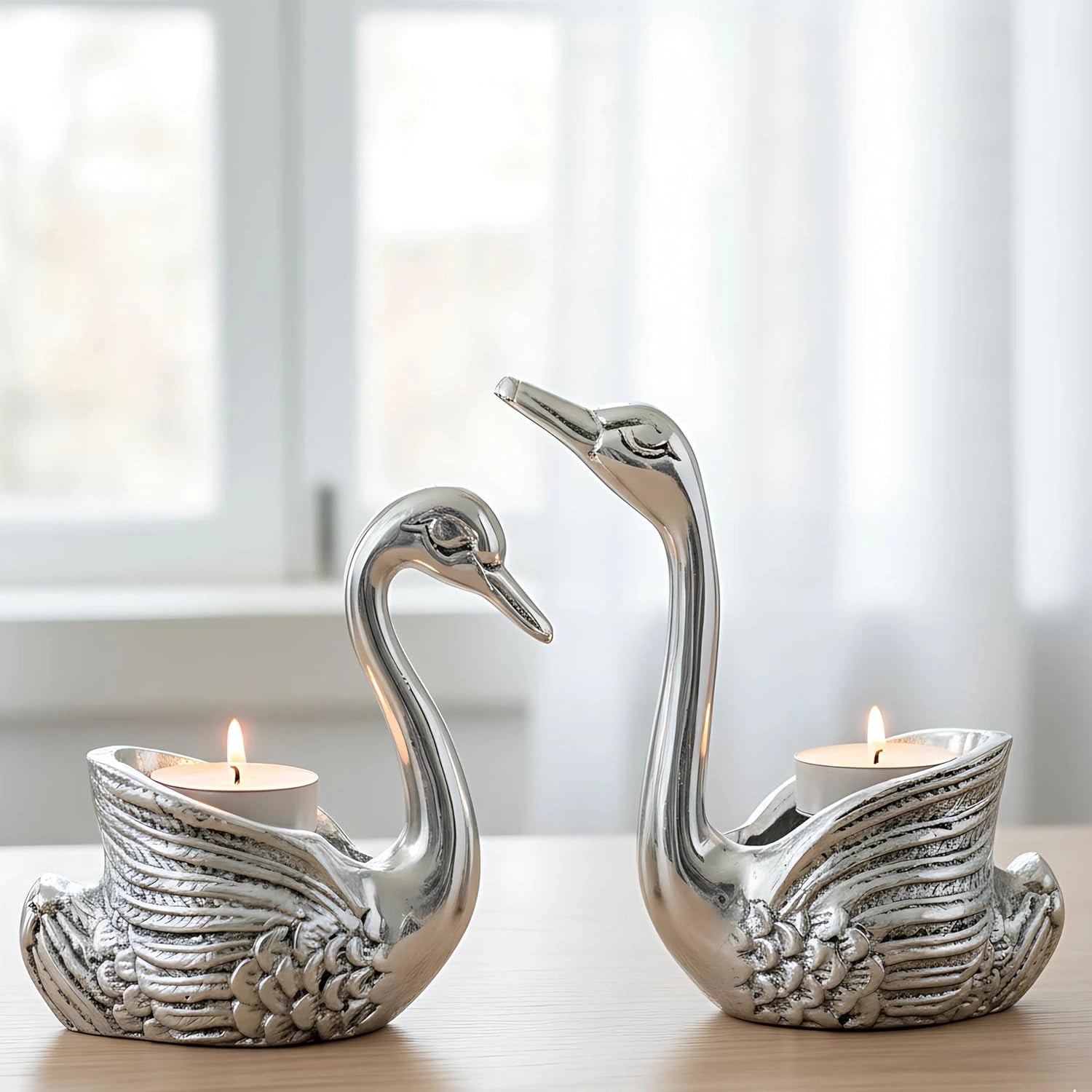 Pair of silver swan-shaped candle holders with lit tea candles on wooden table near window