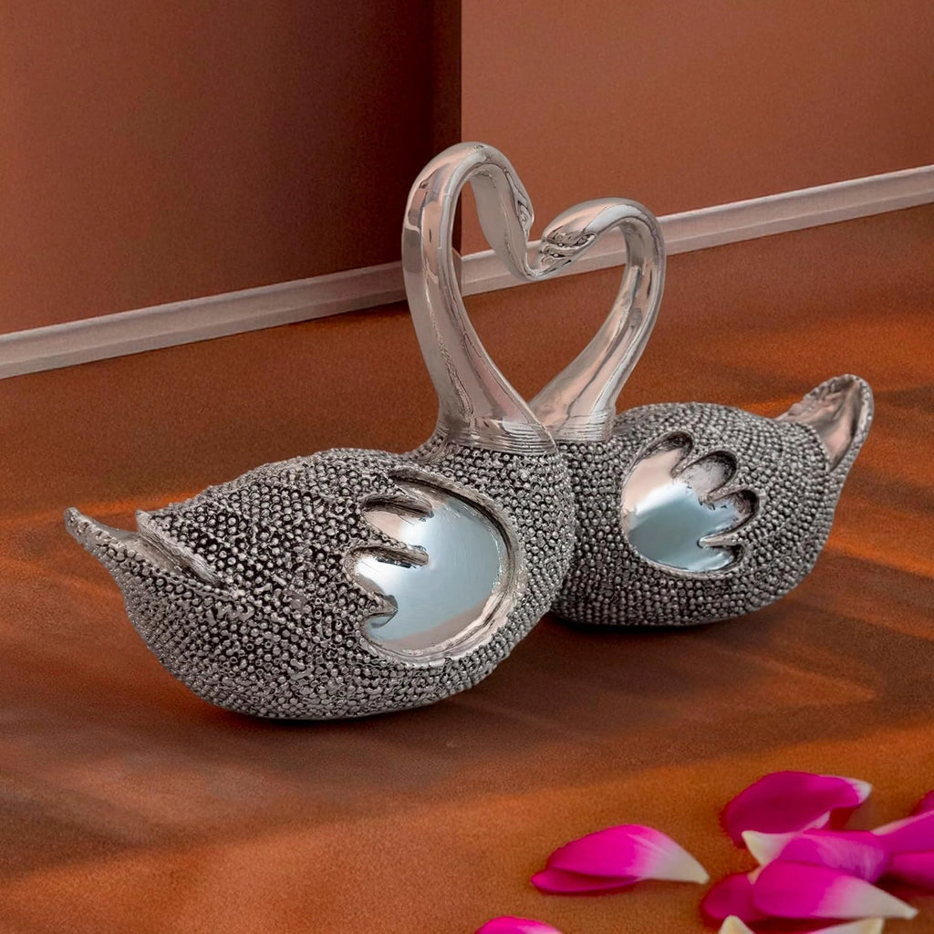 Elegant silver swan figurines with intertwined necks forming a heart on a brown surface with pink petals